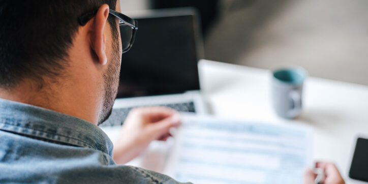 Rear view of male freelancer analyzing business plan over documents while sitting at table in home office.