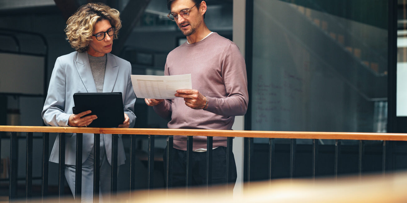 Business people reading a document together. Two professionals analyse a financial report in an office. Business colleagues discuss with each other while standing on an interior balcony.