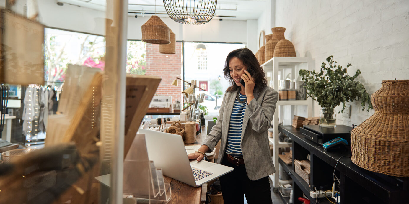 Smiling young Asian woman standing behind a counter in her stylish boutique working on a laptop and talking on a cellphone