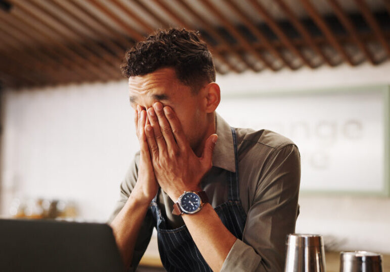 Frustrated man, barista and laptop with mistake for fatigue, eye strain or company bankruptcy at restaurant. Male person, bartender or overworked on computer for fail, loss or debt at indoor bar